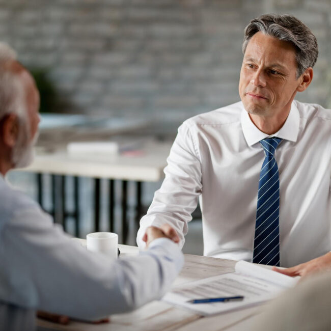Middle-aged businessman in a white shirt and blue striped tie shakes hands with an older man across a desk during a professional meeting, with documents and a pen on the table.