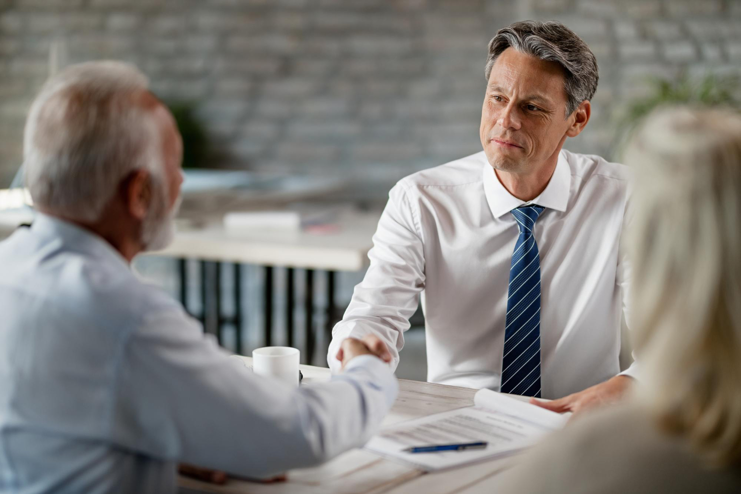 Middle-aged businessman in a white shirt and blue striped tie shakes hands with an older man across a desk during a professional meeting, with documents and a pen on the table.