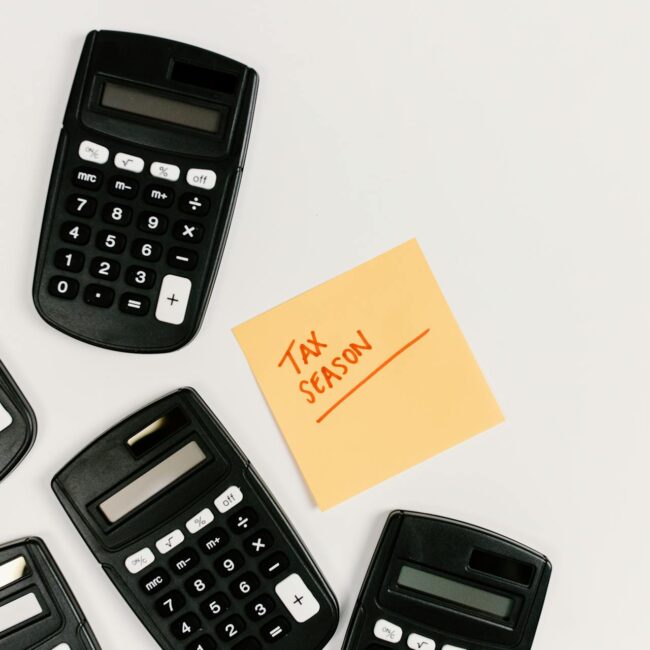 A group of black calculators arranged around a yellow sticky note that reads “TAX SEASON” in red handwriting on a white background.