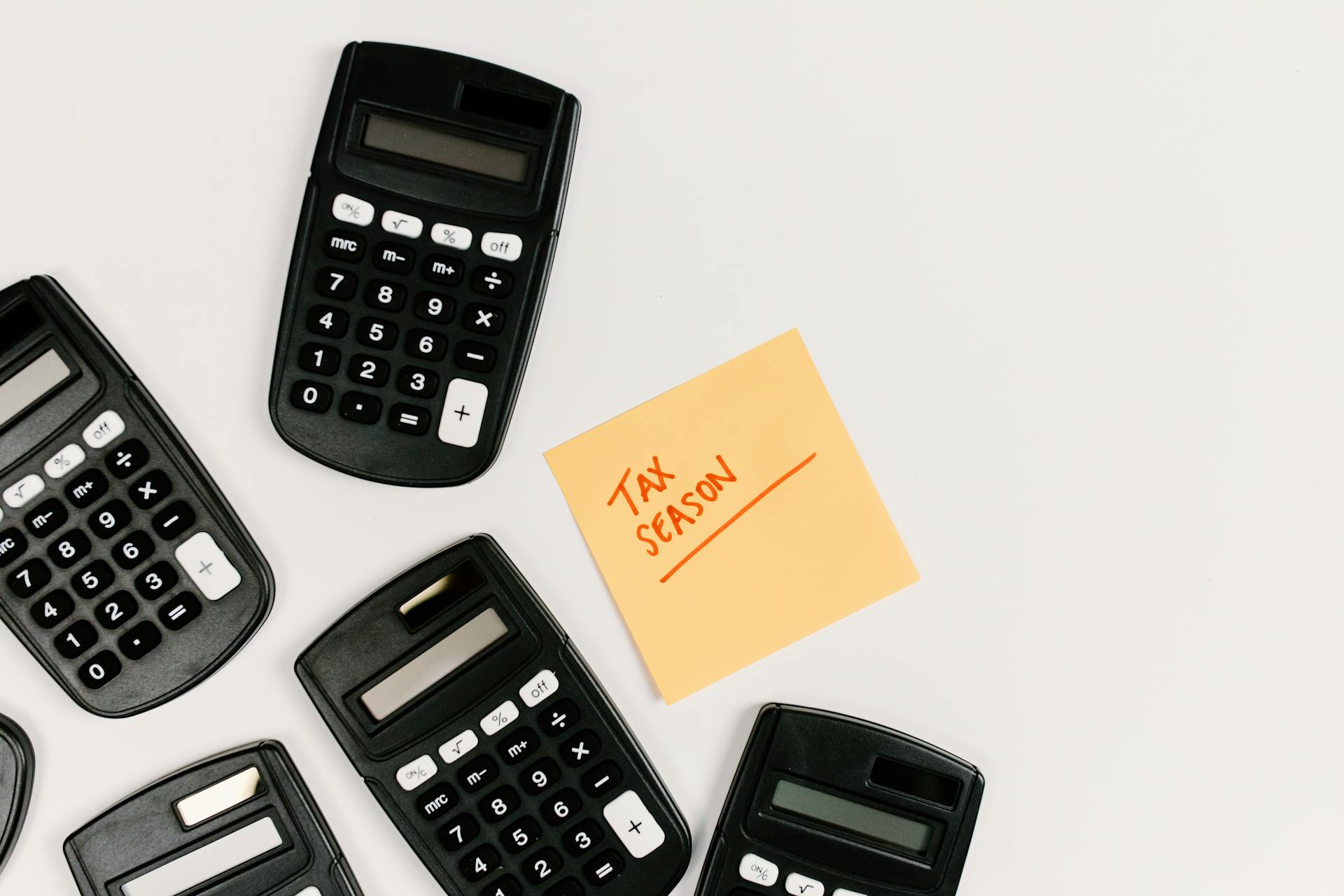 A group of black calculators arranged around a yellow sticky note that reads “TAX SEASON” in red handwriting on a white background.