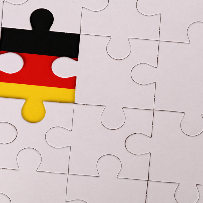 A close-up of a mostly white jigsaw puzzle with one central piece highlighted in the colors of the German flag—black on top, red in the middle, and gold at the bottom—standing out against the surrounding plain pieces.