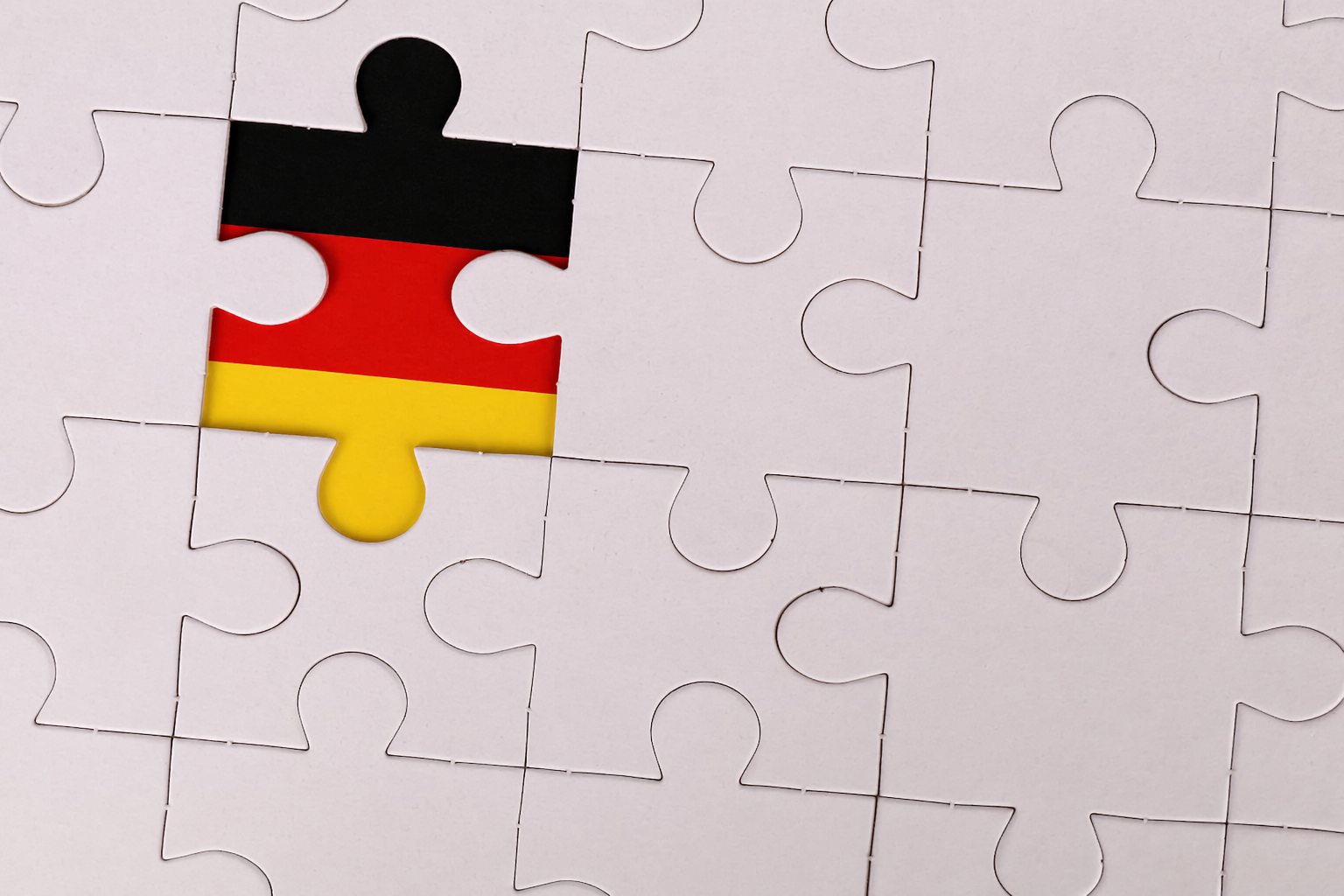 A close-up of a mostly white jigsaw puzzle with one central piece highlighted in the colors of the German flag—black on top, red in the middle, and gold at the bottom—standing out against the surrounding plain pieces.
