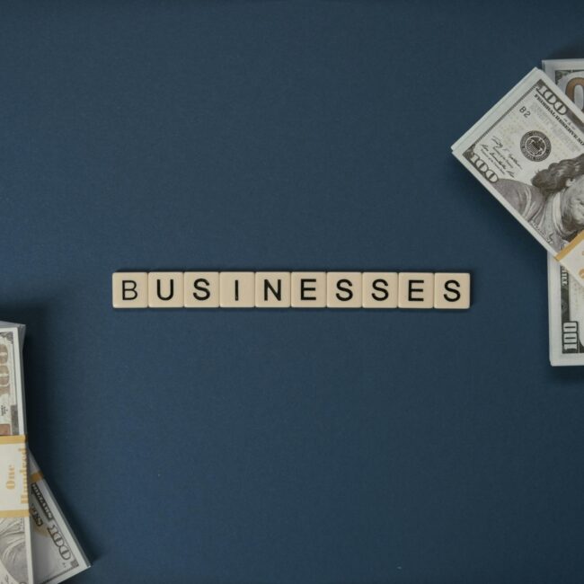 Bundles of US $100 bills placed on both sides of a blue surface, with letter tiles in the center spelling the word “BUSINESSES.”