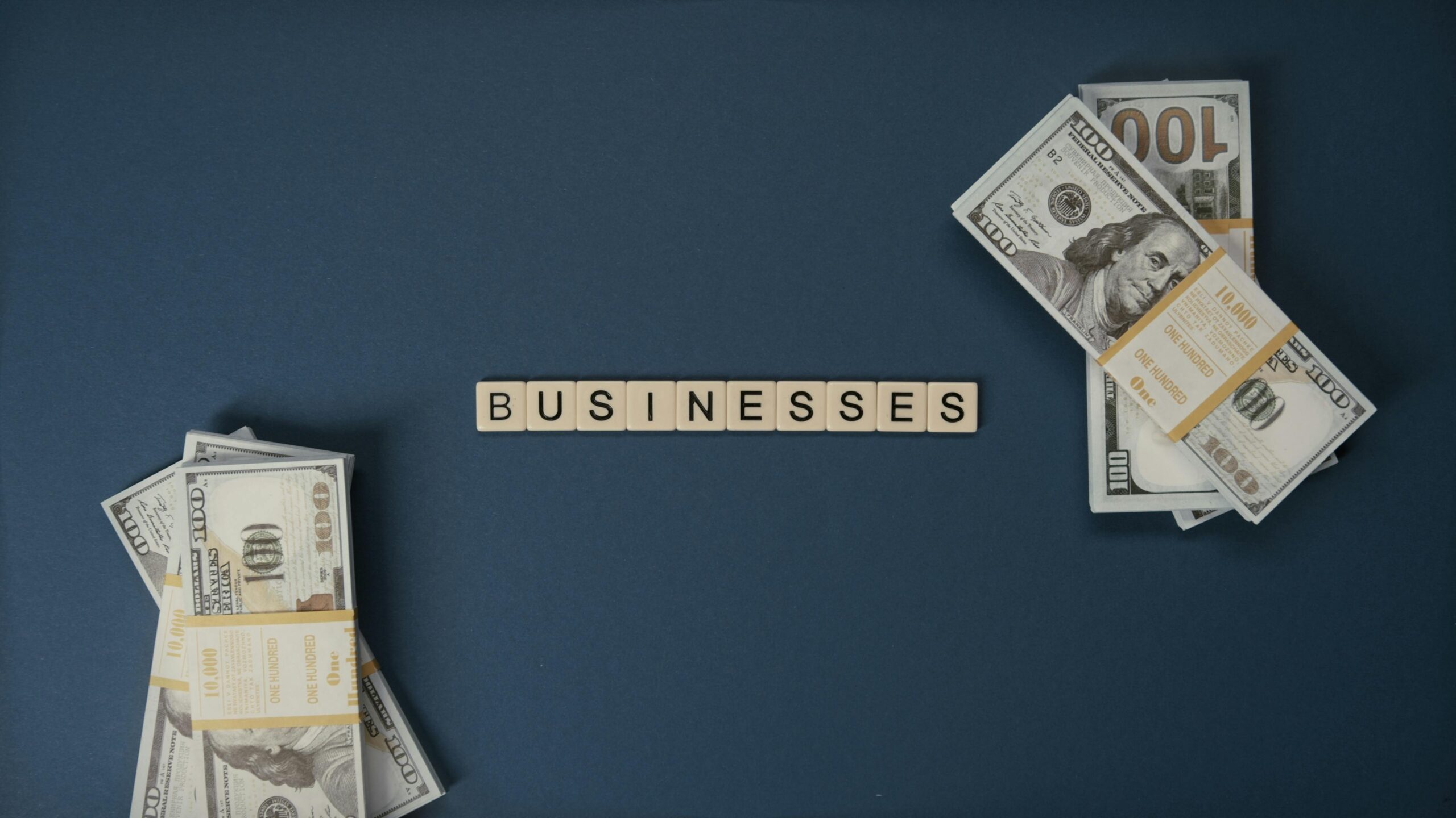 Bundles of US $100 bills placed on both sides of a blue surface, with letter tiles in the center spelling the word “BUSINESSES.”