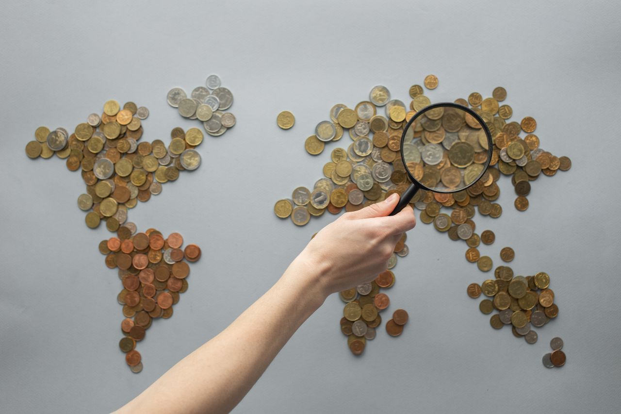 A collection of mixed coins arranged to resemble a world map on a gray surface, with a hand holding a magnifying glass over Europe and Asia, symbolizing global finance, economic analysis, or international markets.