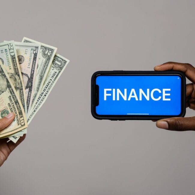 Hands holding U.S. dollar banknotes and a smartphone displaying the word "FINANCE" on a blue screen, illustrating digital finance, payments, and financial management.