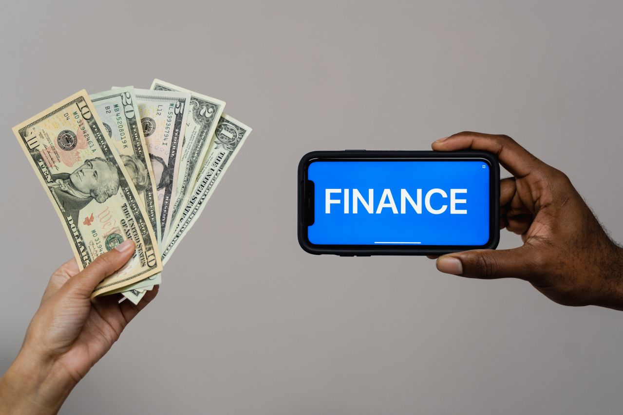Hands holding U.S. dollar banknotes and a smartphone displaying the word "FINANCE" on a blue screen, illustrating digital finance, payments, and financial management.