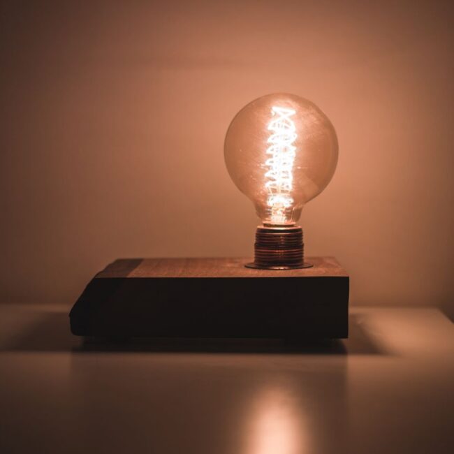 **Alt text:** A glowing vintage-style light bulb sits on a rectangular wooden base in a dimly lit room, casting a warm amber light that softly illuminates the surrounding wall and reflects off the smooth tabletop below.