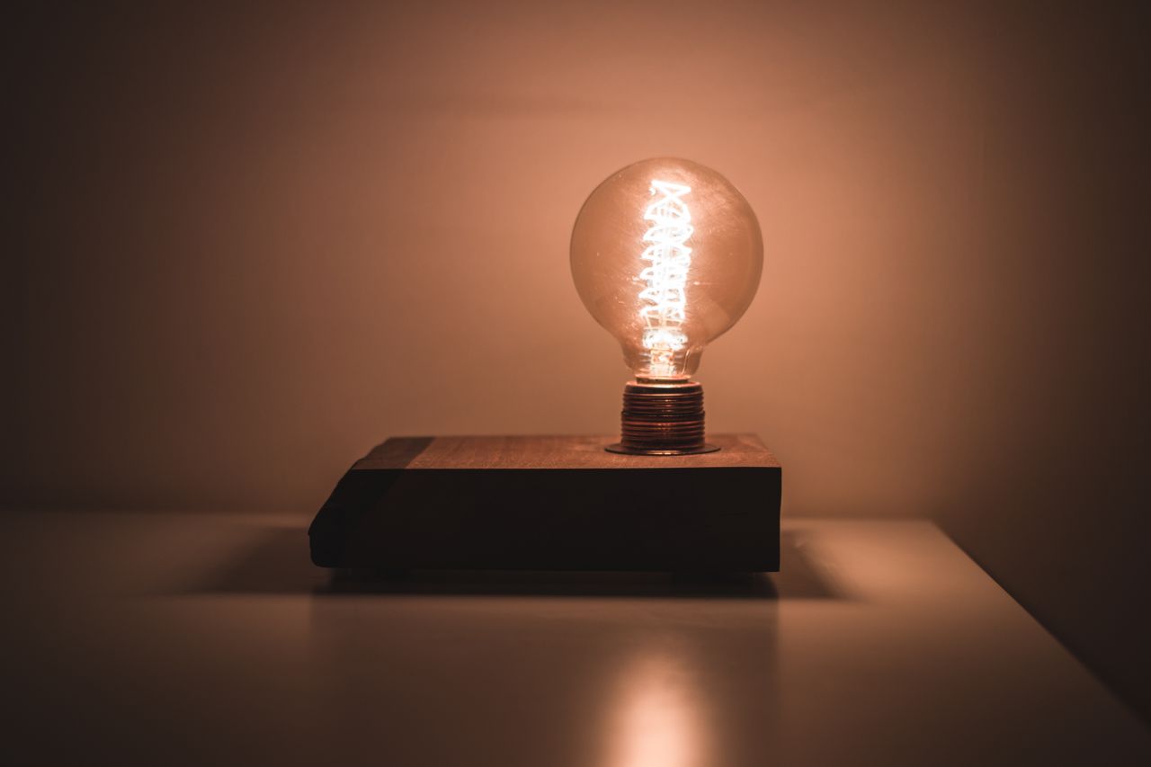 **Alt text:** A glowing vintage-style light bulb sits on a rectangular wooden base in a dimly lit room, casting a warm amber light that softly illuminates the surrounding wall and reflects off the smooth tabletop below.