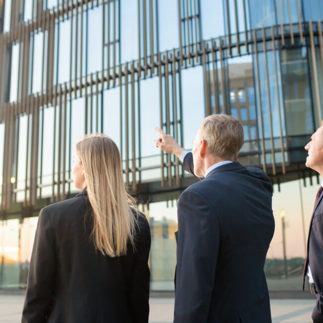 Three business professionals in suits standing outside a modern glass office building, discussing and pointing at the architecture.