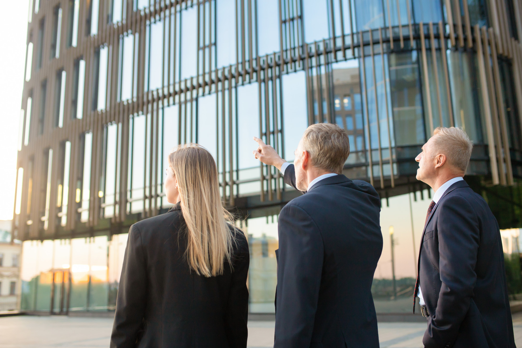 Three business professionals in suits standing outside a modern glass office building, discussing and pointing at the architecture.